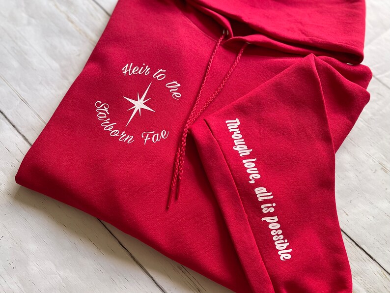 May include: Red hooded sweatshirt with white embroidery. The front reads "Heir to the Starborn Fae" with a star design. The left sleeve reads "Through love, all is possible."