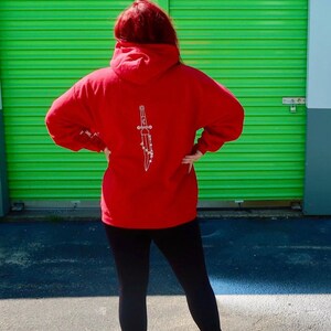 May include: Red hooded sweatshirt with a white graphic of a dagger on the back.