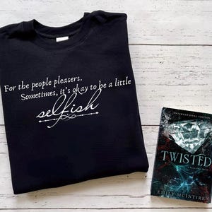 以下が含まれることがあります： 「For the people pleasers. Sometimes, it's okay to be a little selfish」と白い文字で書かれた黒いスウェットシャツ。横には、Emily McIntire著の「Twisted」というタイトルの本があります。