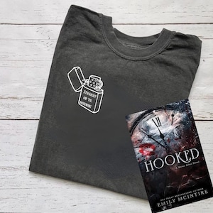 以下が含まれることがあります： 「Straight on til morning」という文字が入ったライターの白いグラフィックが描かれた濃いグレーのTシャツ。背景には、エミリー・マッキンタイア著の「Hooked」というタイトルの本がある。