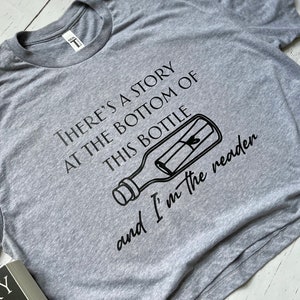 May include: A gray t-shirt with a black graphic of a message in a bottle. The text on the shirt reads "There's a story at the bottom of this bottle and I'm the reader."