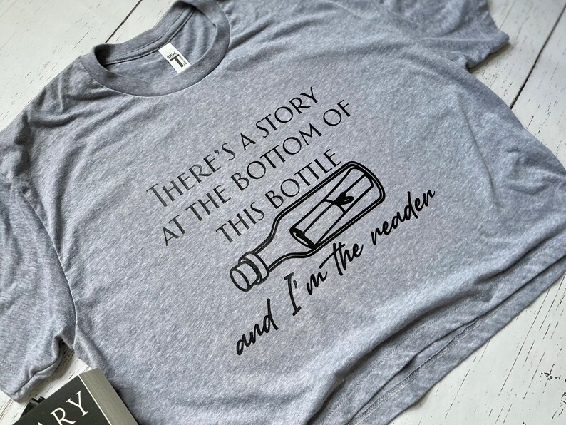 May include: A gray t-shirt with a black graphic of a message in a bottle. The text on the shirt reads "There's a story at the bottom of this bottle and I'm the reader."