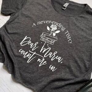 Pu&ograve; includere: T-shirt grigio scuro con testo bianco e grafica di libri e una rosa. Il testo recita: "A never-ending TBR? Dear Maria, count me in."
