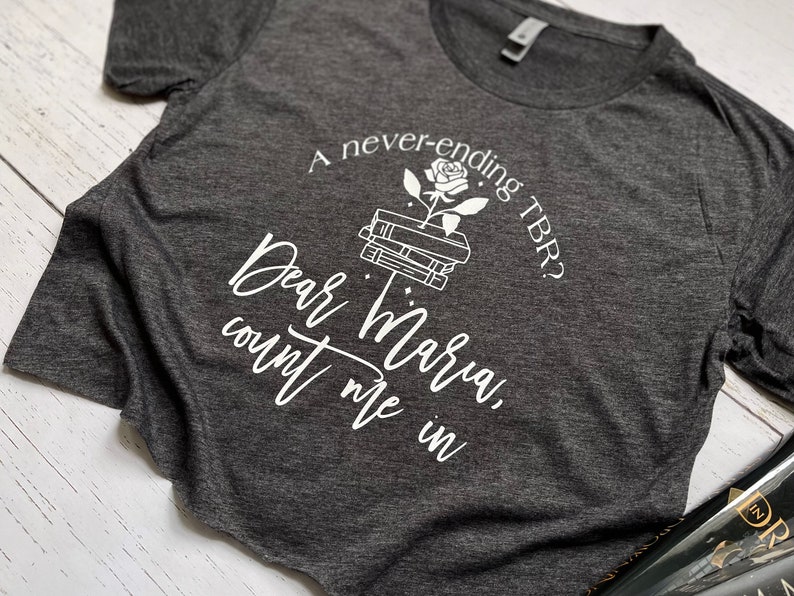 Pu&ograve; includere: T-shirt grigio scuro con testo bianco e grafica di libri e una rosa. Il testo recita: "A never-ending TBR? Dear Maria, count me in."