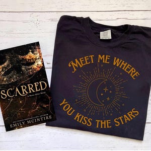以下が含まれることがあります： 白い木製の表面に置かれた黒いTシャツ。Tシャツには、「Meet me where you kiss the stars」という金色のグラフィックと、星がついた三日月が描かれている。Tシャツの隣には、「Scarred」というタイトルのEmily McIntireの本がある。