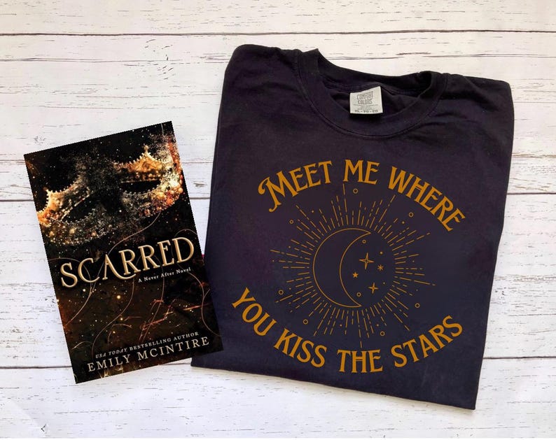 May include: A black t-shirt with a gold graphic that says "Meet me where you kiss the stars" and a crescent moon with stars. The t-shirt is lying on a white wooden surface next to a book titled "Scarred" by Emily McIntire.