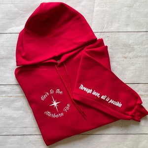 May include: Red hooded sweatshirt with white embroidered text on the front and sleeve. The front text reads "Heir to the Starborn Fire" with a white star in the center. The sleeve text reads "Through love, all is possible".