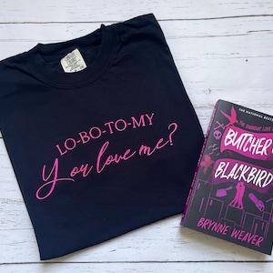 以下が含まれることがあります： ピンクの文字で「LO-BO-TO-MY You love me?」と書かれた黒いTシャツ。Tシャツの隣には、ブリン・ウィーバー著の「ブッチャー＆ブラックバード」というタイトルのペーパーバックがあります。