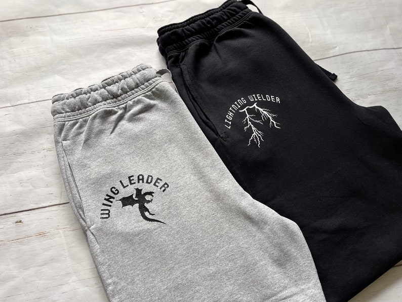 May include: Two pairs of sweatpants, one gray and one black, with drawstring waistbands and side pockets. The gray sweatpants have a black dragon graphic and the text "WING LEADER". The black sweatpants have a lightning bolt graphic and the text "LIGHTNING WIELDER".