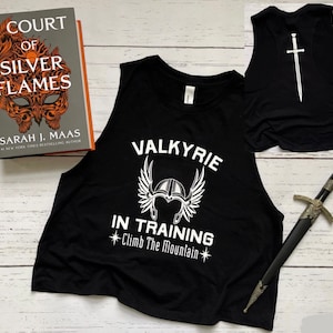 May include: Black sleeveless crop top with white text that reads "Valkyrie in Training *Climb The Mountain*" and a white graphic of a winged helmet. The back of the shirt has a white sword graphic.