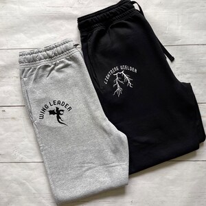 May include: Two pairs of sweatpants, one gray and one black, are displayed on a light wood surface. The gray sweatpants have the text "WING LEADER" with a graphic. The black sweatpants have the text "LIGHTNING WELDER" with a lightning bolt graphic.
