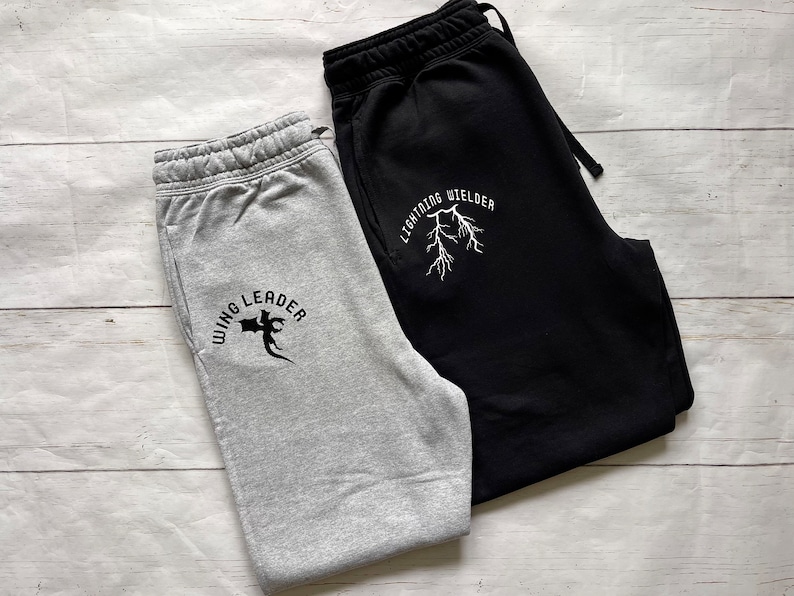 May include: Two pairs of sweatpants, one gray and one black, are displayed on a light wood surface. The gray sweatpants have the text "WING LEADER" with a graphic. The black sweatpants have the text "LIGHTNING WELDER" with a lightning bolt graphic.