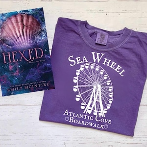 以下が含まれることがあります： 「Sea Wheel Atlantic Cove Boardwalk」という文字と白い観覧車のグラフィックが描かれた紫色のTシャツ。Tシャツの隣には、表紙に貝殻が描かれた「Hexed」というタイトルの本がある。