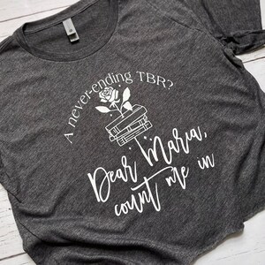 Pu&ograve; includere: T-shirt grigio scuro con testo bianco e grafica di una rosa che cresce da una pila di libri. Il testo recita: "A never-ending TBR? Dear Maria, count me in."