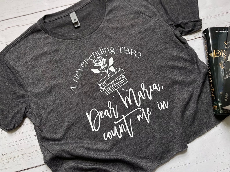 Pu&ograve; includere: T-shirt grigio scuro con testo bianco e grafica di una rosa che cresce da una pila di libri. Il testo recita: "A never-ending TBR? Dear Maria, count me in."
