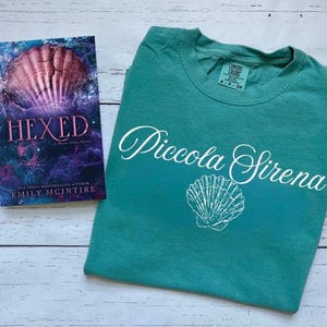 以下が含まれることがあります： 白い木製の表面に置かれた、ターコイズブルーのTシャツ。白い筆記体で「Piccola Sirena」という文字と、白い貝殻のグラフィックが描かれている。Tシャツの隣には、エミリー・マッキンタイア著「Hexed」というタイトルの本がある。