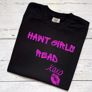 May include: A black short-sleeved t-shirt featuring bright pink text that reads HAWT GIRLS READ XOXO, accompanied by a pink graphic of a pair of lips. The shirt has a crew neckline and a visible brand label on the inside of the collar.
