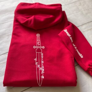 May include: Red hooded sweatshirt with a white outline of a sword with a crescent moon and the word "QUINLAN" on the back. The left sleeve has the text "Through love, all is possible" in white.