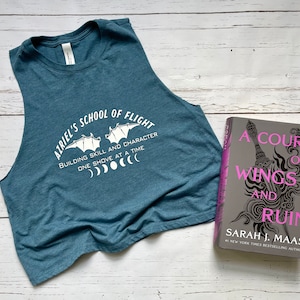 Może przedstawiać: Niebieska koszulka na ramiączkach z napisem "Azriel's School of Flight" i grafiką dwóch nietoperzy. Tekst pod nietoperzami brzmi "Building skill and character one shove at a time". Koszulka leży obok książki zatytułowanej "A Court of Wings and Ruin" autorstwa Sarah J. Maas.