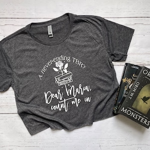 Pu&ograve; includere: T-shirt grigio scuro con testo bianco: "A never-ending TBR? Dear Maria, count me in." Una grafica di rosa e libri &egrave; sopra il testo. Tre libri sono visibili a destra.