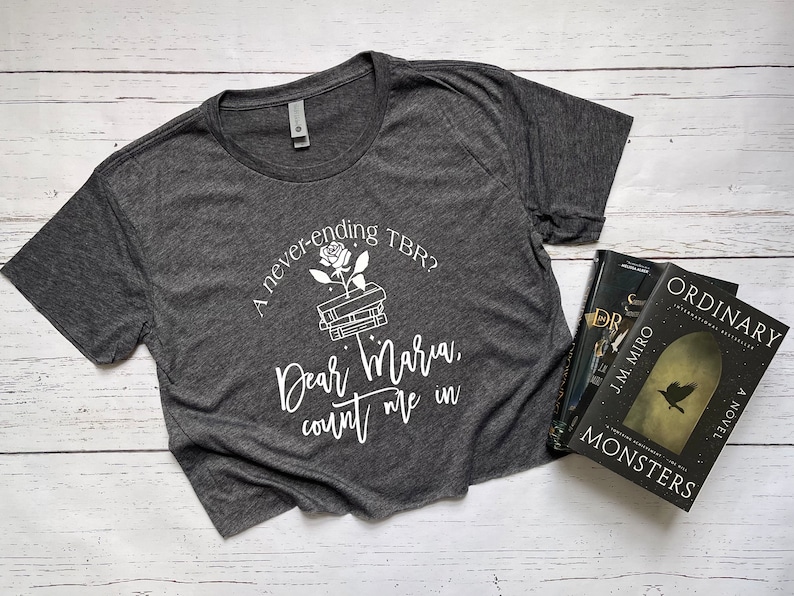 Pu&ograve; includere: T-shirt grigio scuro con testo bianco: "A never-ending TBR? Dear Maria, count me in." Una grafica di rosa e libri &egrave; sopra il testo. Tre libri sono visibili a destra.