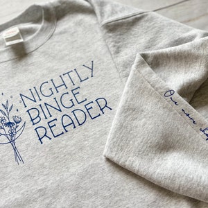 Könnte beinhalten: Ein grauer Pullover mit dem Text "Nightly Binge Reader" und einem Halbmond mit Blumen, der auf der Vorderseite aufgedruckt ist. Der Ärmel hat den Text "One more chapter..." aufgedruckt.