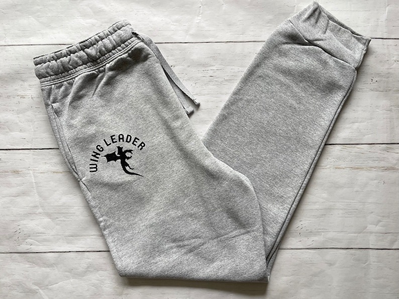 May include: Light gray sweatpants with a drawstring waist and elastic cuffs. The left leg features a black graphic with the words "WING LEADER" in a semi-circular design above a stylized dragon.