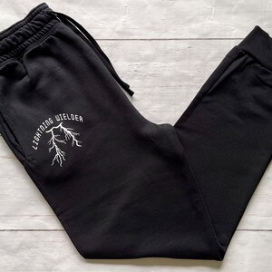 May include: Black sweatpants with a drawstring waist and cuffed ankles. The left leg features the white text "LIGHTNING WIELDER" above a white lightning bolt graphic. The pants are made of a soft material and are suitable for casual wear.