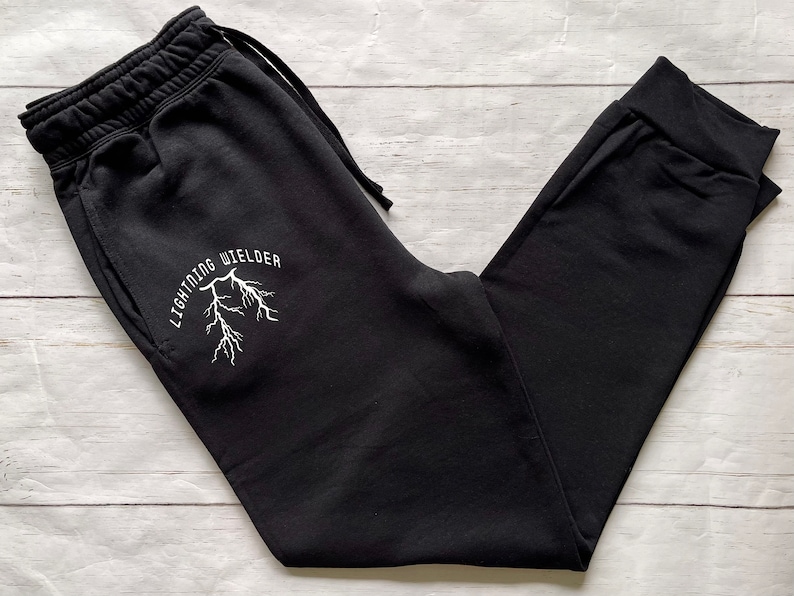 May include: Black sweatpants with a drawstring waist and cuffed ankles. The left leg features the white text "LIGHTNING WIELDER" above a white lightning bolt graphic. The pants are made of a soft material and are suitable for casual wear.