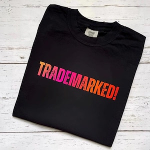 May include: A black short-sleeved t-shirt featuring the word TRADEMARKED! printed in a distressed, gradient font that transitions from pink to orange. The garment has a crew neckline and a visible interior brand label at the collar area.