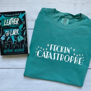 以下が含まれることがあります： 白い文字で「FECKIN' CATASTROPHE」とプリントされたターコイズブルーのTシャツ。Tシャツは、ブリン・ウィーバー著の「Leather & Lark」というタイトルのペーパーバックの隣に、白い木製の表面の上に置かれています。