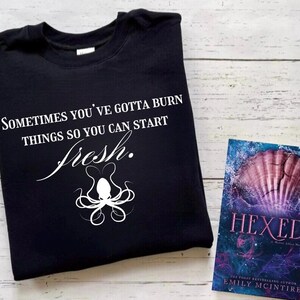 以下が含まれることがあります： 白い文字で「Sometimes you've gotta burn things so you can start fresh.」と書かれた黒いTシャツ。テキストの下には白いタコのデザインがあります。背景には「Hexed」というタイトルの本があります。