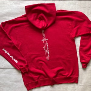 May include: Red hooded sweatshirt with a white outline of a dagger with the text "Through love, all is possible" on the left sleeve and "QUINLAN" written on the dagger.