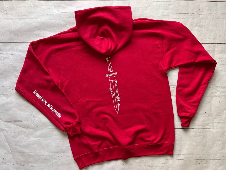 May include: Red hooded sweatshirt with a white outline of a dagger with the text "Through love, all is possible" on the left sleeve and "QUINLAN" written on the dagger.