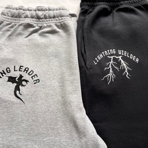 May include: Two pairs of sweatpants, one gray and one black. The gray sweatpants have the text "WING LEADER" with a dragon graphic. The black sweatpants have the text "LIGHTNING WIELDER" with a lightning bolt graphic.