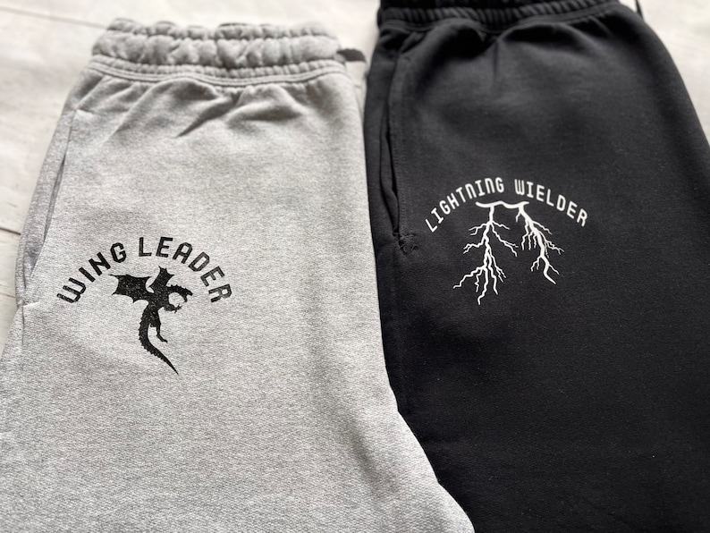 May include: Two pairs of sweatpants, one gray and one black. The gray sweatpants have the text "WING LEADER" with a dragon graphic. The black sweatpants have the text "LIGHTNING WIELDER" with a lightning bolt graphic.