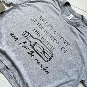 May include: Gray t-shirt with a black graphic of a bottle with a message inside. The text on the shirt reads "There's a story at the bottom of this bottle and I'm the reader."