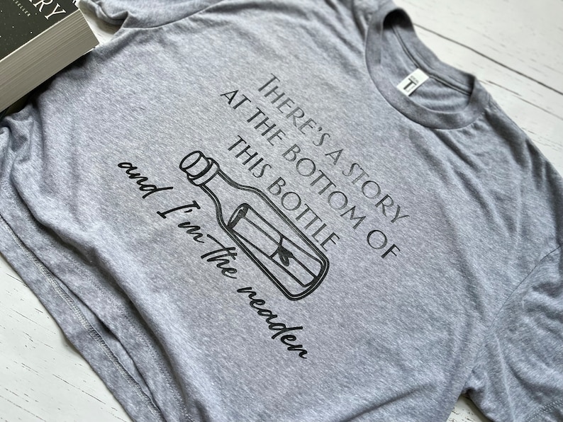 May include: Gray t-shirt with a black graphic of a bottle with a message inside. The text on the shirt reads "There's a story at the bottom of this bottle and I'm the reader."
