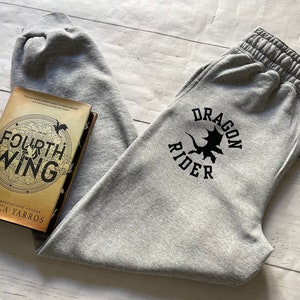 May include: Gray sweatpants with a black "Dragon Rider" logo featuring a dragon silhouette. The sweatpants are lying on a wooden surface next to a book titled "The Fourth Wing" by Rebecca Yarros.