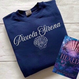 以下が含まれることがあります： 白い文字で「Piccola Sirena」と書かれたネイビーブルーのスウェットシャツ。スウェットシャツには貝殻のグラフィックもプリントされています。背景にはエミリー・マッキンタイアの「Hexed」というタイトルの本があります。