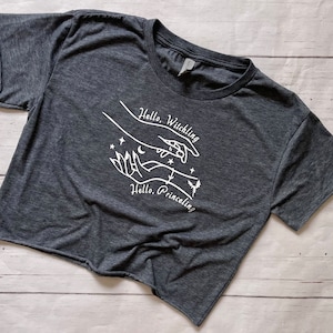 May include: A dark gray cropped t-shirt with a white graphic design of two hands reaching towards each other. The design includes a crescent moon, stars, and the text "Hello, Witchling" and "Hello, Princeeling".