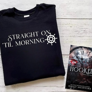 以下が含まれることがあります： 白い文字で「ストレートオン 'Til モーニング」と白い船のホイールのグラフィックが書かれた黒いTシャツ。Tシャツは、「フックド」というタイトルの時計の文字盤のグラフィックが描かれた本の隣にあります。