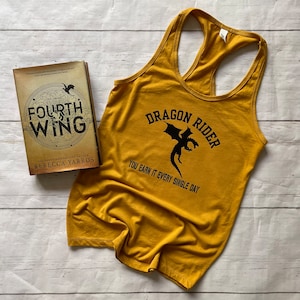 Peut inclure: Un débardeur jaune moutarde avec un motif de dragon noir et le texte "Dragon Rider You earn it every single day". Le débardeur est à côté d'un livre intitulé "Fourth Wing" de Rebecca Yarros.