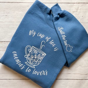 Könnte beinhalten: Ein blaues Sweatshirt mit weißem Text, der "My cup of tea is enemies to lovers" lautet, und einer weißen Illustration einer Teetasse mit aufsteigendem Dampf. Der rechte Ärmel hat weißen Text, der "Spill the tea" lautet, und eine weiße Illustration einer Teekanne.