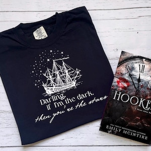 以下が含まれることがあります： 星が輝く夜空の下を航海する船の白いグラフィックがプリントされたネイビーブルーのTシャツ。Tシャツには「Darling, if I'm the dark, then you're the stars」というテキストがプリントされています。Tシャツは、エミリー・マッキンタイア著の「Hooked」というタイトルの本の隣にあります。