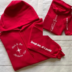 May include: Red hooded sweatshirt with white text and a star design on the front. The text reads "Heir to the Starborn Five". The sleeve has the text "Through love, all is possible".