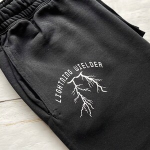 May include: Black sweatpants with an elastic waistband and a pocket. The white text "LIGHTNING WIELDER" arches above a white lightning bolt graphic. The pants are made of a soft material.