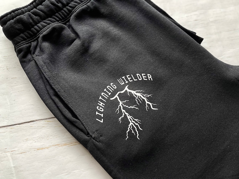May include: Black sweatpants with an elastic waistband and a pocket. The white text "LIGHTNING WIELDER" arches above a white lightning bolt graphic. The pants are made of a soft material.