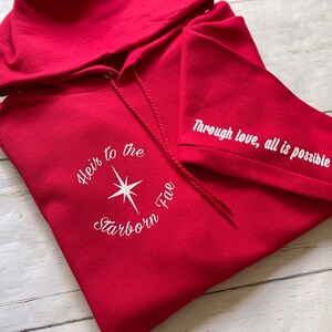 May include: Red hooded sweatshirt with white embroidered text "Heir to the Starborn Fire" on the front and "Through love, all is possible" on the sleeve.