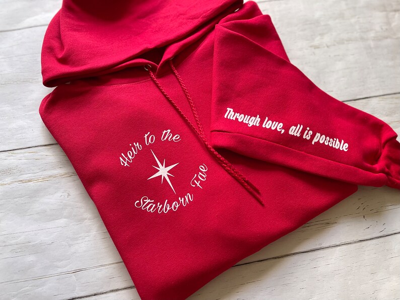 May include: Red hooded sweatshirt with white embroidered text "Heir to the Starborn Fire" on the front and "Through love, all is possible" on the sleeve.